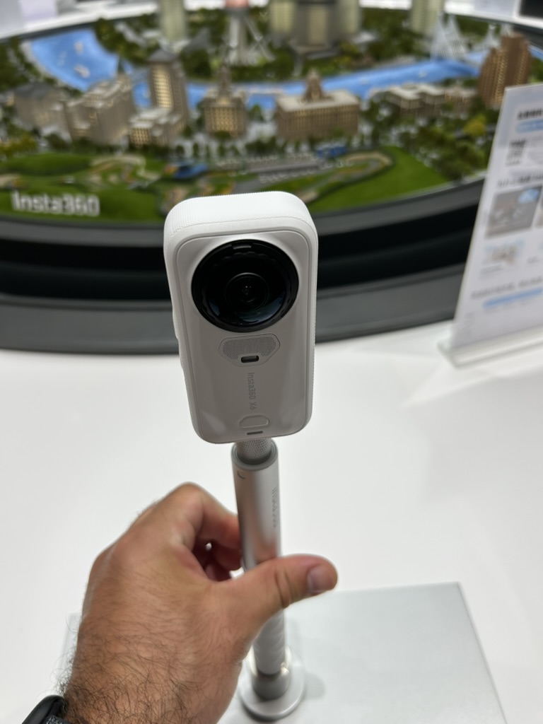 White Insta360 X6 on a selfie stick, held in front of a miniature city display